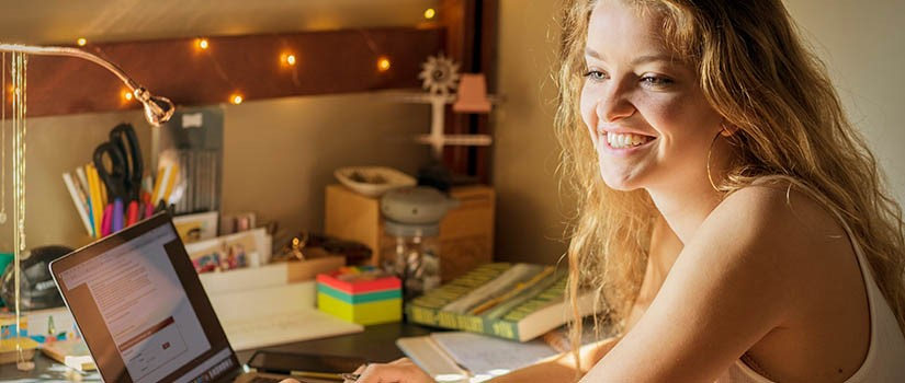 a white female student sits at a desk looking at her computer. She is smiling.