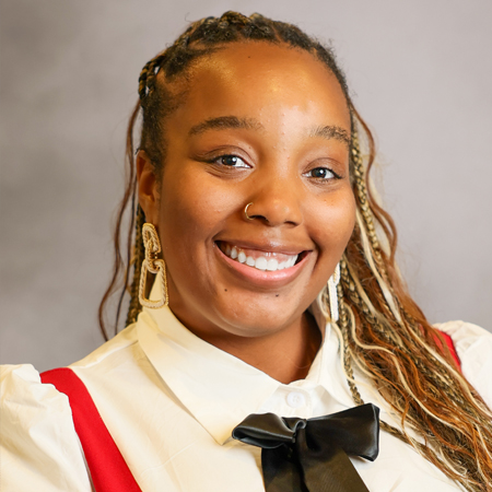 Professional headshot of a smiling person with long braided hair featuring blonde highlights wearing a white collared blouse with a black bow and a red sleeveless dress. The person is facing the camera and is wearing a small nose ring and large decorative earrings. The photo is set against a soft gray studio background with warm, even lighting, creating a friendly, approachable, and professional appearance.