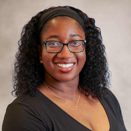 Professional headshot of a smiling person with shoulder-length curly black hair held back with a black headband, wearing black rectangular eyeglasses and a fitted black short-sleeve top. The person is facing the camera and is wearing small stud earrings and a delicate gold necklace with a geometric pendant. The photo is set against a soft gray studio background with warm, even lighting, creating a friendly, approachable, and professional appearance.