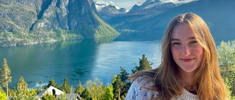 Student smiling in foreground with long hair, wearing a patterned sweater, standing above a scenic fjord with mountains, water and blue sky with clouds in the background.