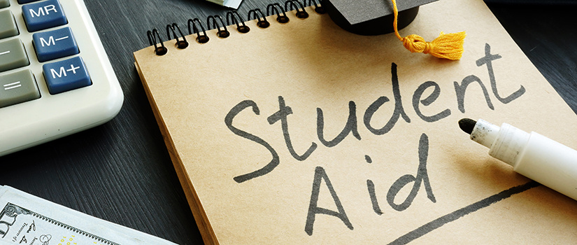 A notepad with the words Student Aid written on top along with a small graduation cap. A calculator and paper money can also be seen on the edges.